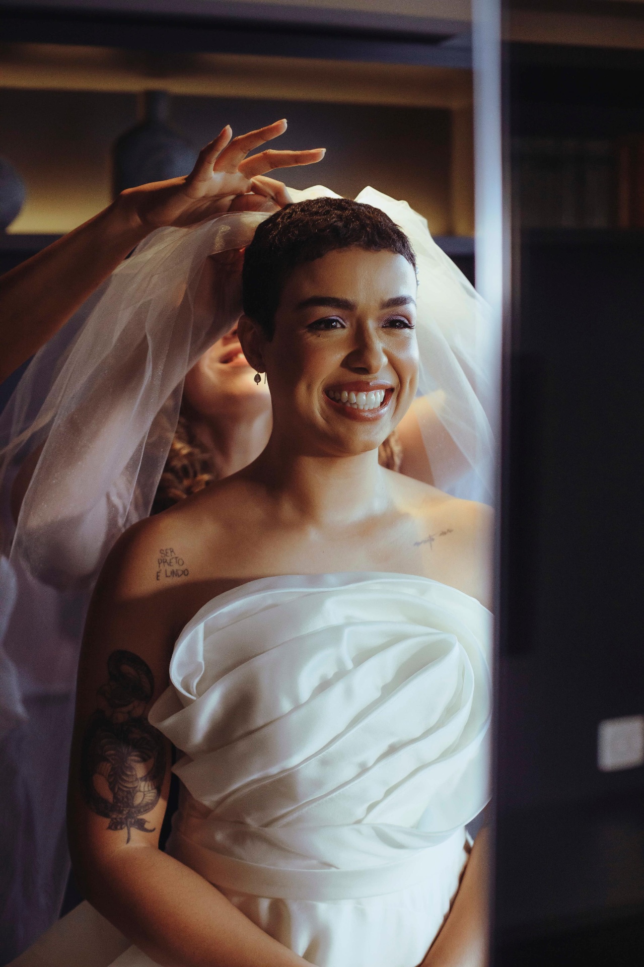 Noiva de cabelo curto sorrindo enquanto ajustam o véu